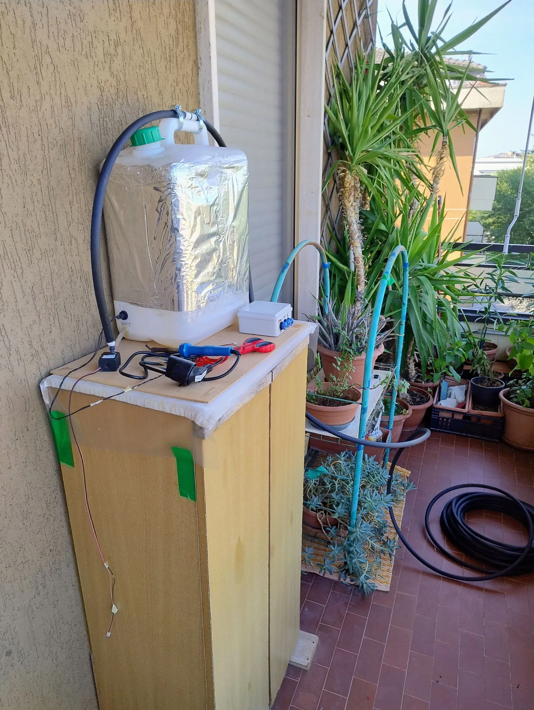 The system on the balcony: a white tank wrapped in aluminium foil on top of a wooden cabinet, a box with the controller and blue connectors, and black hoses running to the pots