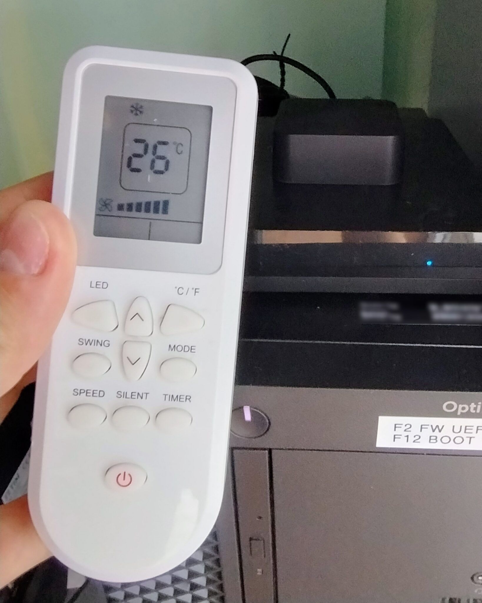 A hand holds a white air-conditioner remote showing 26 degrees in front of a black PC with a small dark cube on top