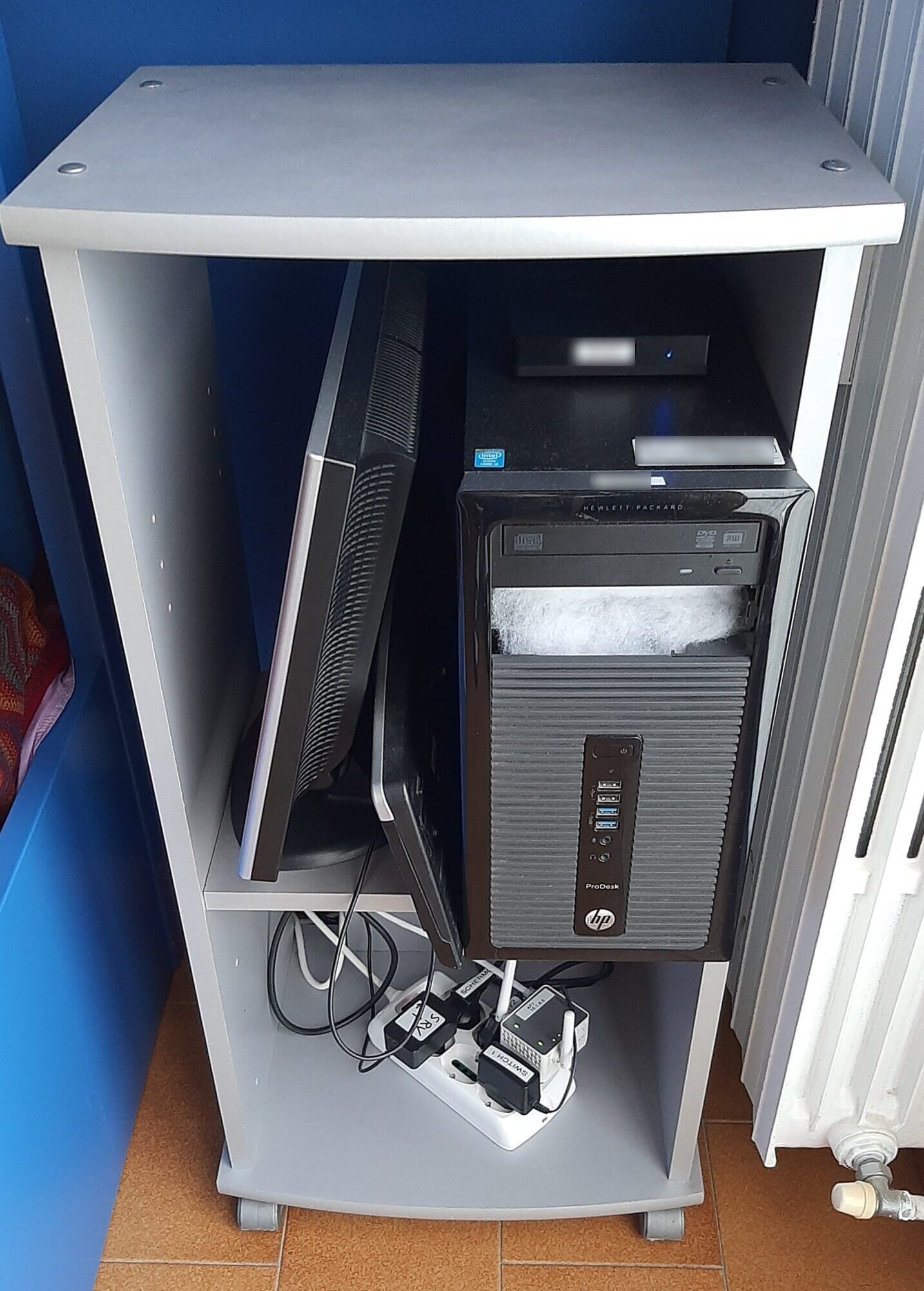 A grey cupboard holding a black HP PC, a monitor leaning on its side and, at the bottom, a power strip with adapters