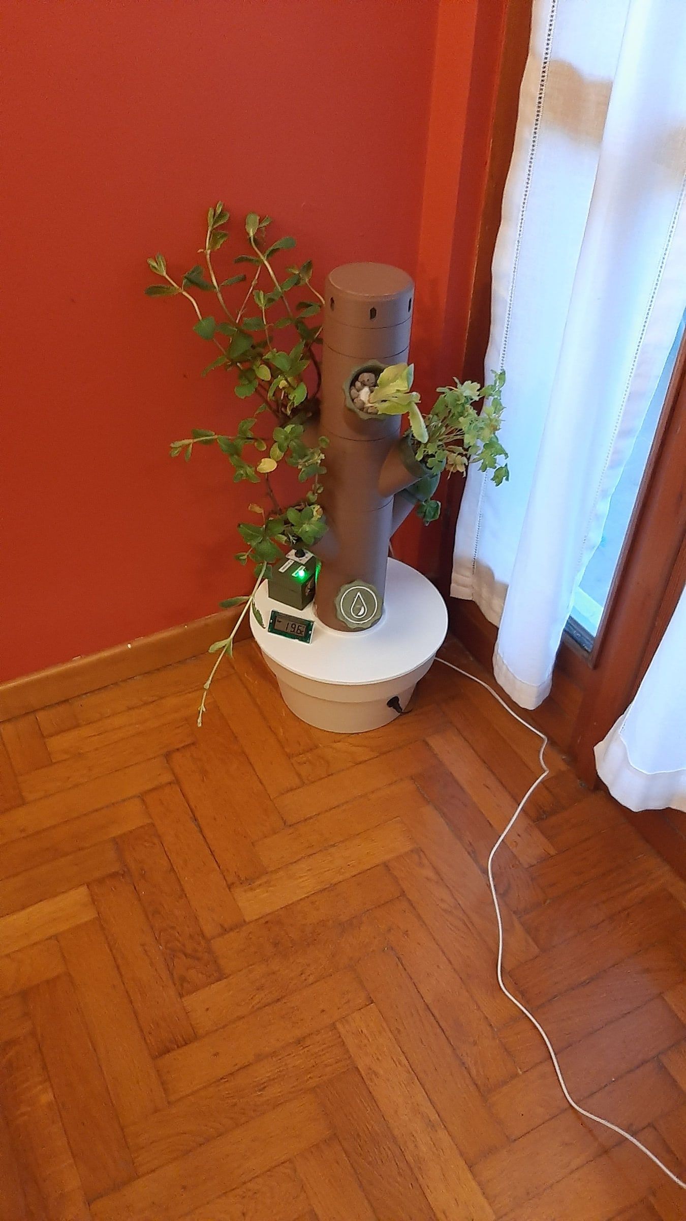 A brown 3D-printed tower with green plants coming out of its holes, standing on a white pot in a corner of a room with a red wall and a parquet floor