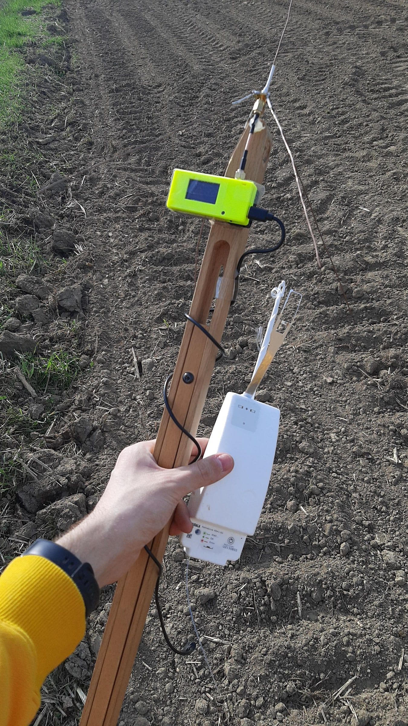 A white radiosonde just recovered, tied to a stick together with the yellow receiver, above a ploughed field