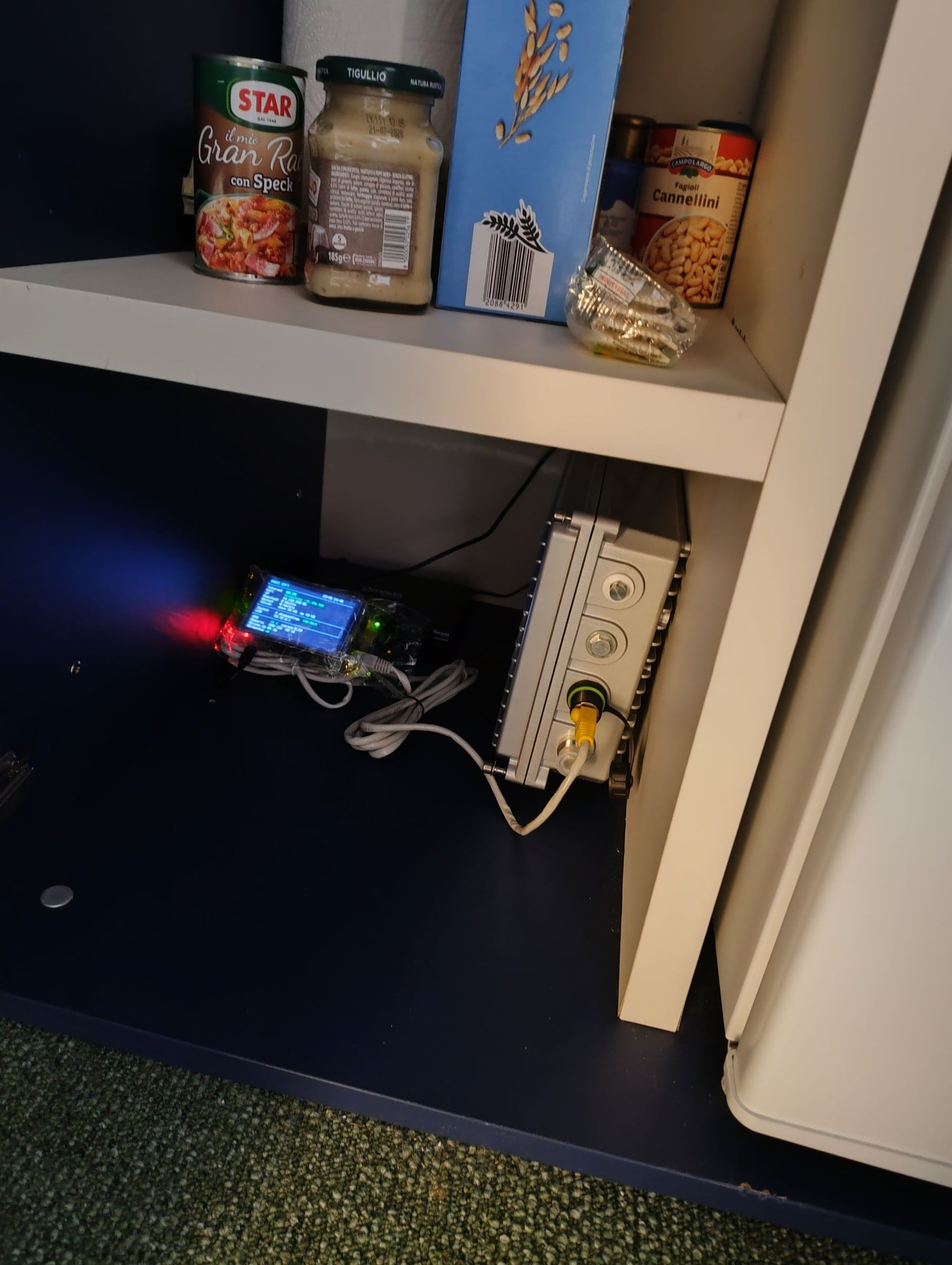 A Raspberry Pi with a small lit screen and white cables, in a dark pantry, next to a grey box with cable glands