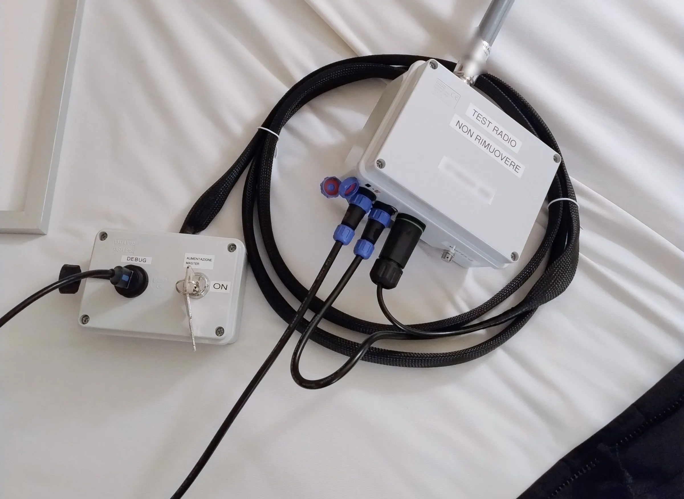 A closed grey gateway with its antenna and black cables, lying on a bed next to a smaller box with a socket and a key switch