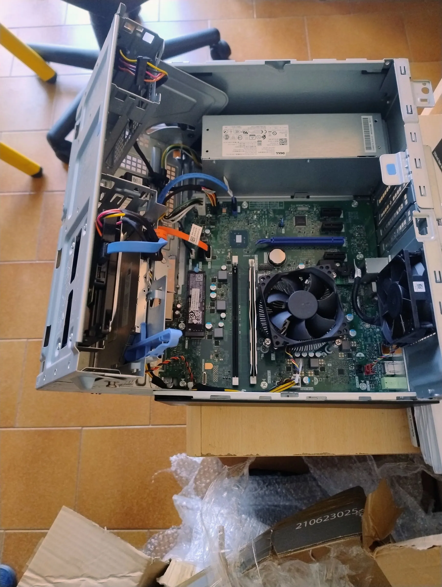 Un PC Dell aperto su un pavimento di piastrelle, con la scheda madre, il dissipatore e l’alimentatore in vista, accanto all’imballaggio
