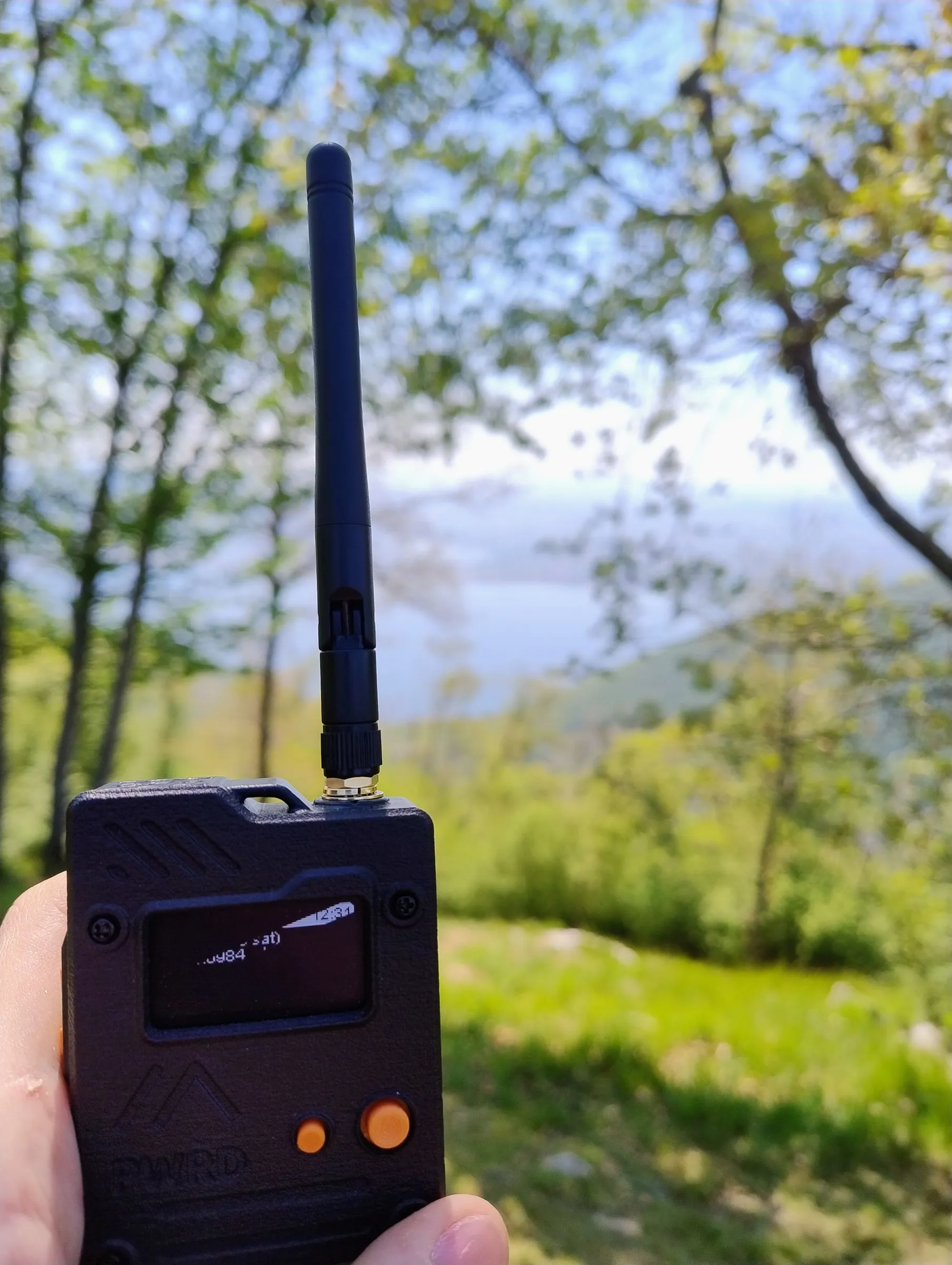 An EnjoyBuds device held in a hand, with a lake and mountains in the background