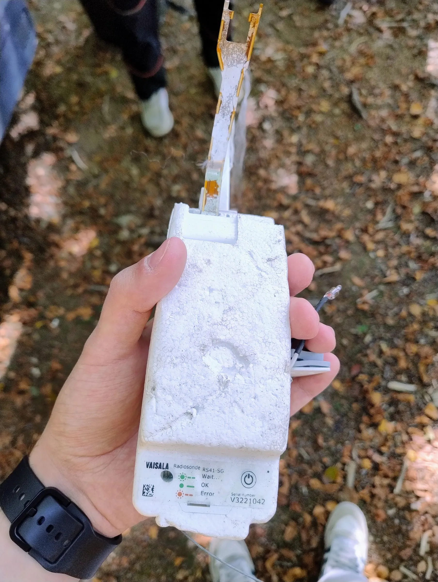 A Vaisala RS41 radiosonde held in a hand in the woods, with the label and serial number in view